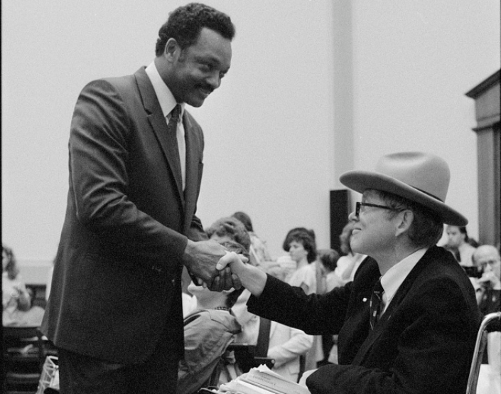 A photo taken in the chambers of the US House of Representatives. Justin Dart, a white man who is sitting in a wheelchair and wearing a suit and a cowboy hat, shakes hands with Reverend Jesse Jackson, an African American man with a mustache who is also wearing a suit. 