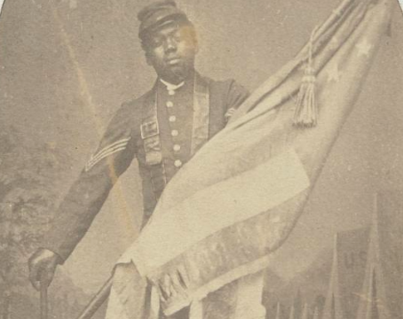 A Black Union soldier in a sergeant's uniform proudly cradles the United States flag that he carried at Fort Wagner. He uses a cane. He stands in front of a false background of army tents. 