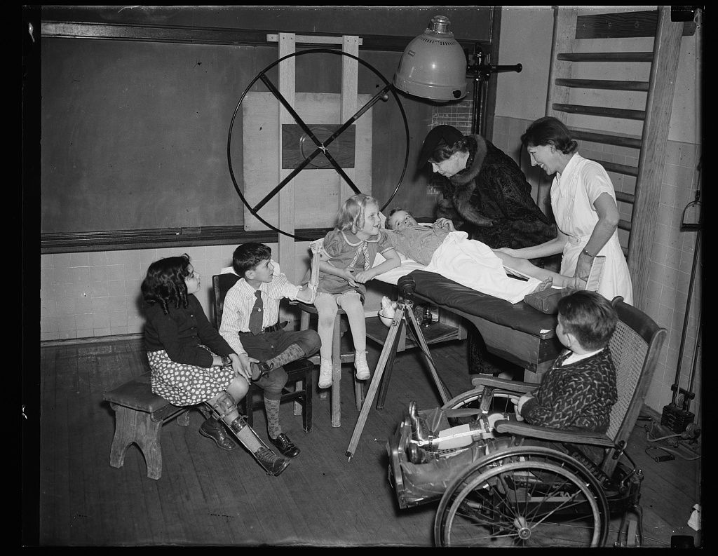 A classroom in a children's school with a blackboard and equipment in the background. A group of children are sitting or standing in front of a medical table, and one boy is laying down in front of the table. A nurse in a white uniform leans over the boy, adjusting his leg. Eleanor Roosevelt, wearing a large fur coat, smiles down at the boy from behind the table. The children have cheerful expressions. 