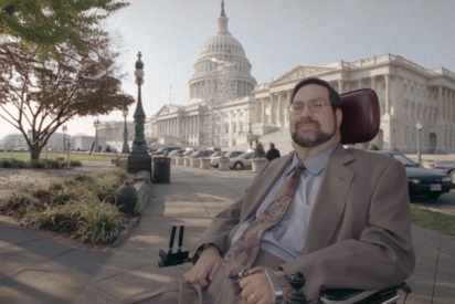 A photograph of activist Tony Young on a street in Washington, D.C. with the Capitol Dome in the background. Young is wearing a suit and uses a motorized wheelchair. 