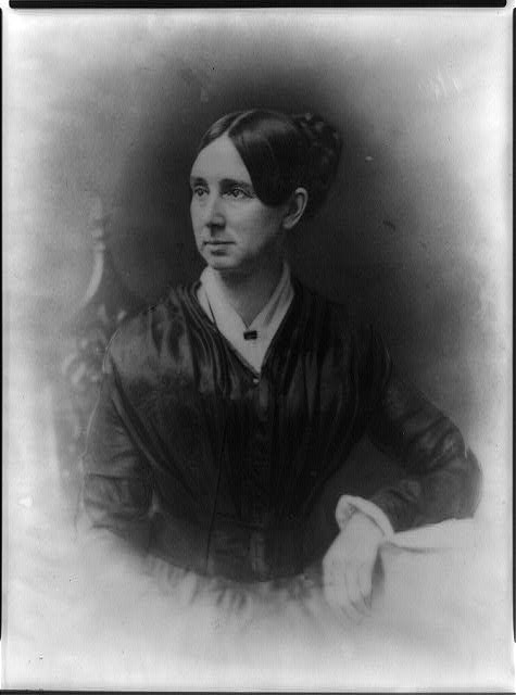 A portrait of Dorothea Lynde Dix, a middle aged woman with dark hair swept back from her face. She wears a formal dress and undershirt. She sits on a wooden chair with ornate scrollwork and rests her arm on a table. She looks to her right with a calm expression. 