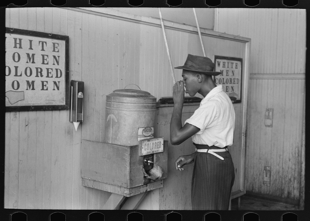 At a tin water dispenser labeled "Colored" a young black man drinks from a paper cup.