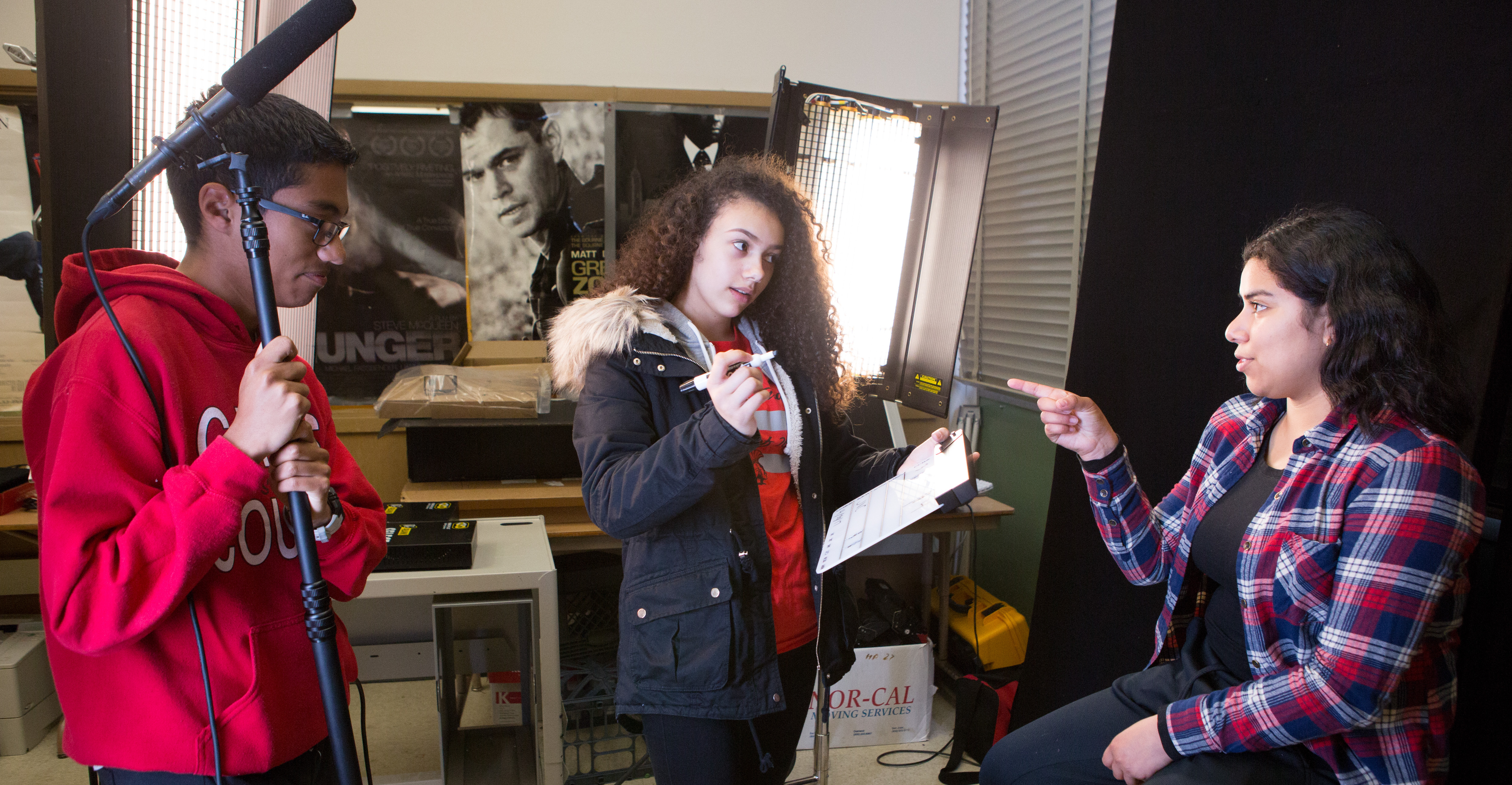 A teenaged girl with long tightly curly hair holds a pen and clipboard. A boy with short black hair and dark skin holds a boom microphone. They are interviewing a girl with a plaid shirt and shoulder length black wavy hair, who makes a point with her hand as she speaks. 