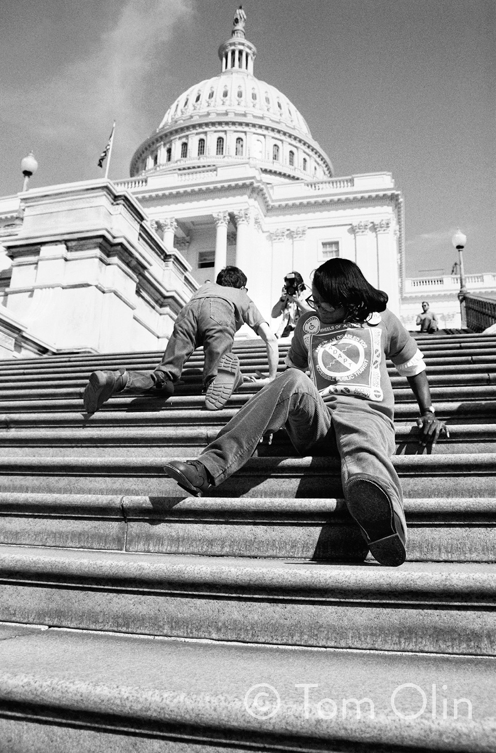 The steps of U.S. Capitol Building with the dome in the background. The camera is aimed up the stairs, as activists crawl up the steps, some of them carrying crutches. It shows how inaccessible the Capitol is. They are protesting in favor of the Americans With Disabilities Act. 