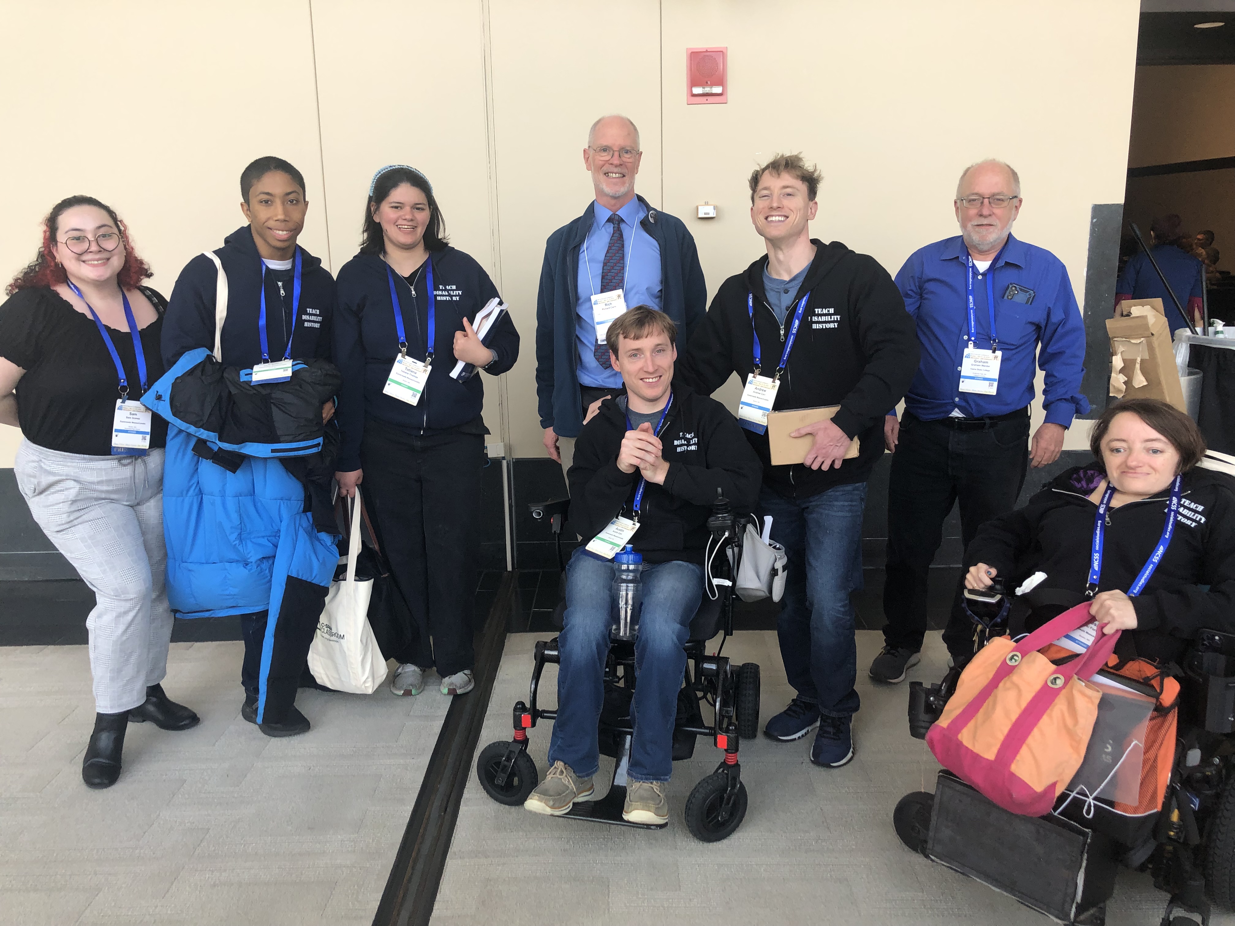 A diverse group of disability activists smile and pose. Five wear Teach Disability History sweatshirts. Two use wheelchairs. All wear conference badges.