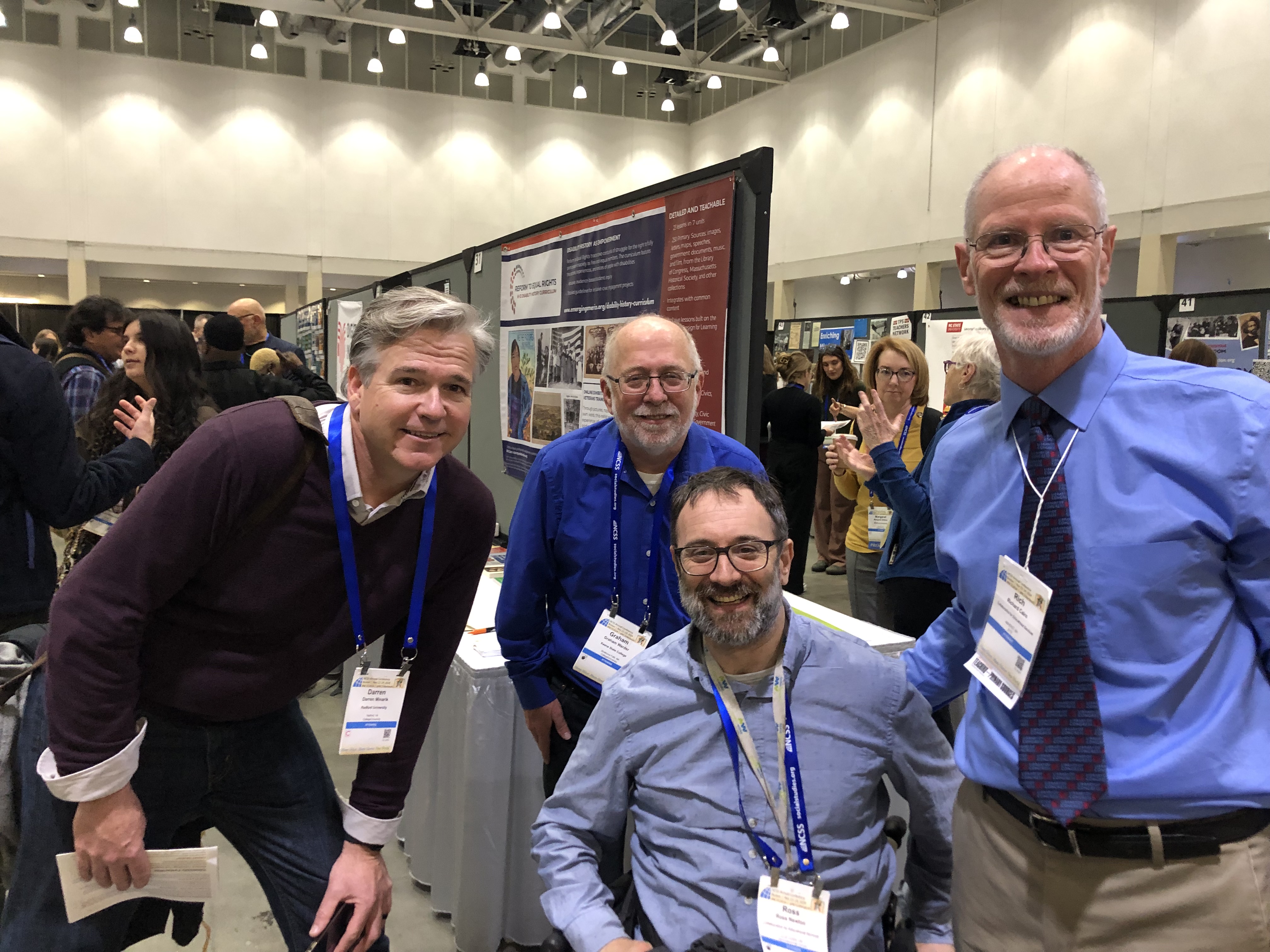 Three men stand and one uses his wheelchair posing in front of a conference poster on teaching disability history. 