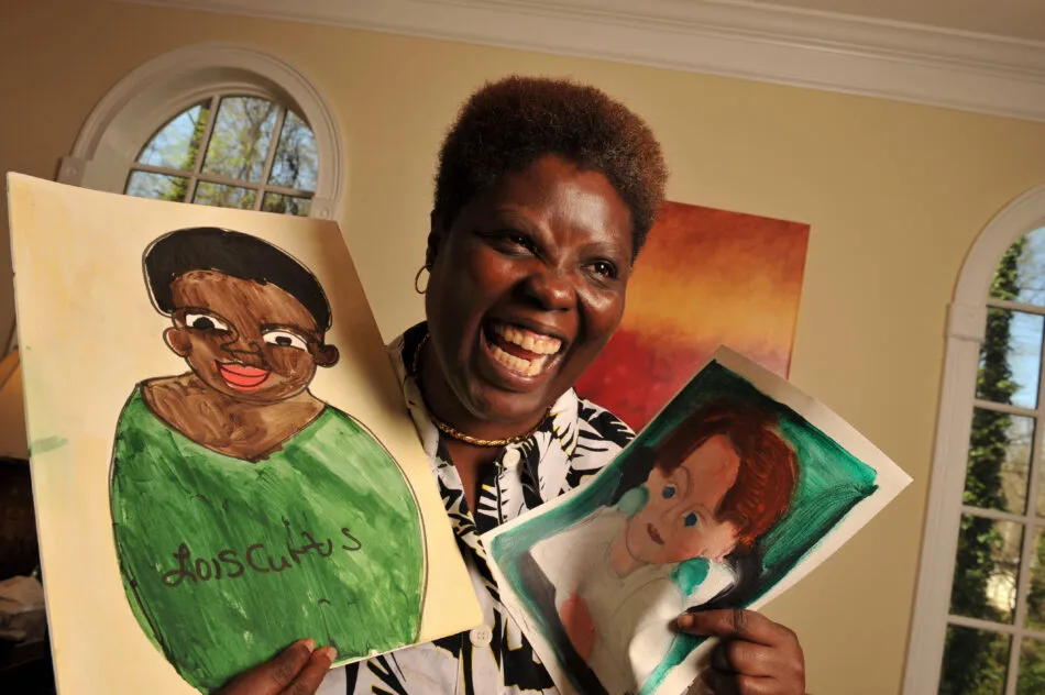 A black woman, Lois Curtis smiles brillilantly as she holds up two of her paintings, one of her and one of her lawyer in the Olmstead v. L.C. case. She is in the Oval Office. 