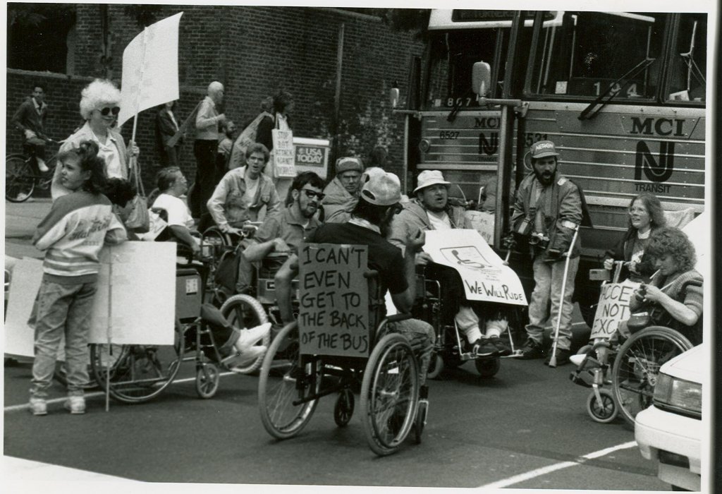 A group of ADAPT Activists protest for accessible public transportation in the street outside a transit center. The activists are mostly wheelchair users. They hold or wear handmade signs, including one saying, "I can't even get to the back of the bus."