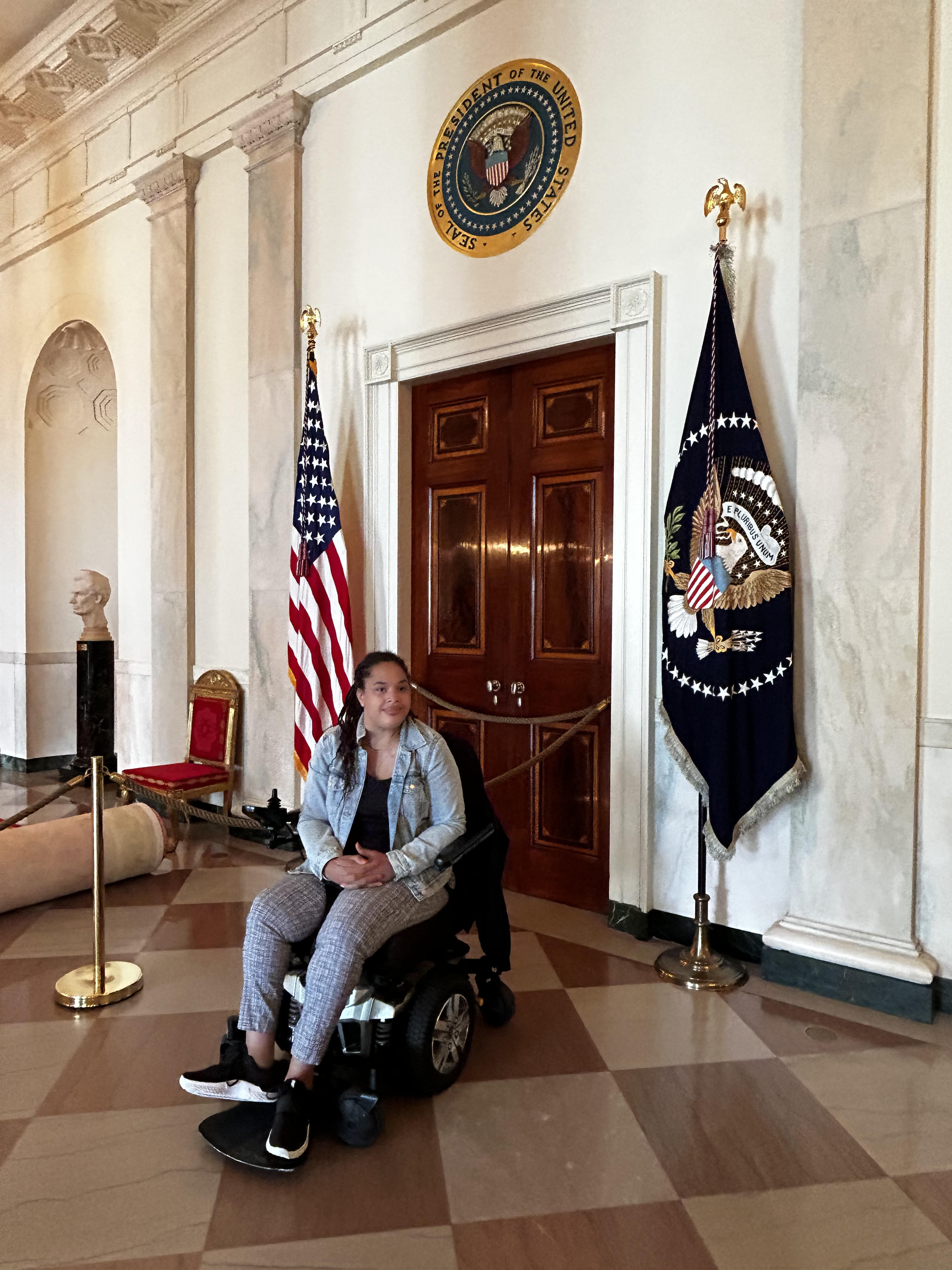 A Black woman using a wheelchair wearing a jeans jacket and with long braids poses in the U.S. White House - a U.S. flag and the flag with the seal of the U.S. President are behind her