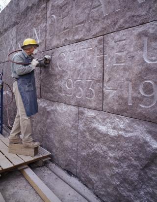 Craftsperson standing on scaffold next to stone wall, protected by yellow hardhat, respirator mask, and apron, etches large letters with a power tool.