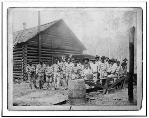 Two dozen black men in striped prison uniforms and hats pose in front of a log cabin