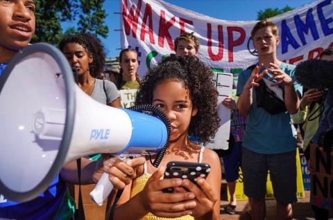 "A young black girl speaks into a bullhorn held by a black teen - a diverse group of teens stands behind them - a banner reads "Wake Up America""