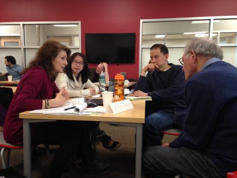 A white-haired man with glasses listens intently as he sits at a table with a diverse group of teachers