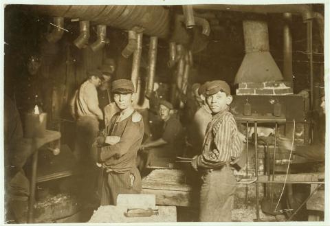 2 boys in ragged clothes and overalls, in cluttered workplace, fireplace and more than 10 tin exhaust pipes in background.