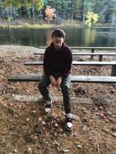 A European American boy with medium-length hair and glasses wears sneakers and a sweatshirt as he sits on bench by a wooded lake in fall